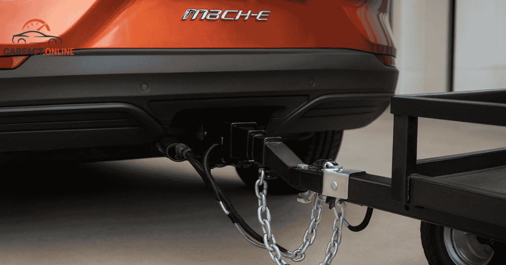 Rear view of a Ford Mustang Mach-E showing trailer hitch, wiring harness, and safety chains connected to a small trailer.